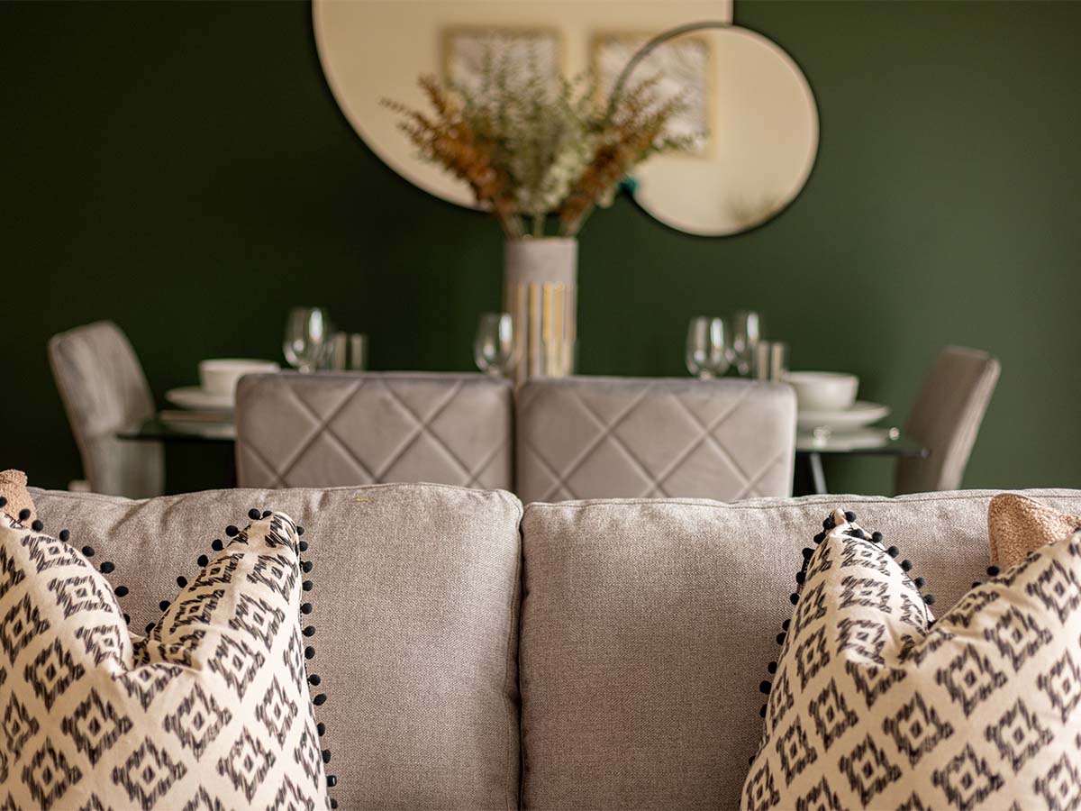 A view from behind a sofa with patterned cushions, looking towards a sophisticated dining area set against a dark green feature wall