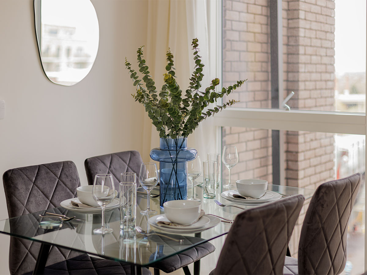 A modern glass dining table set for four with grey quilted velvet chairs and a blue vase of eucalyptus, overlooking a city view