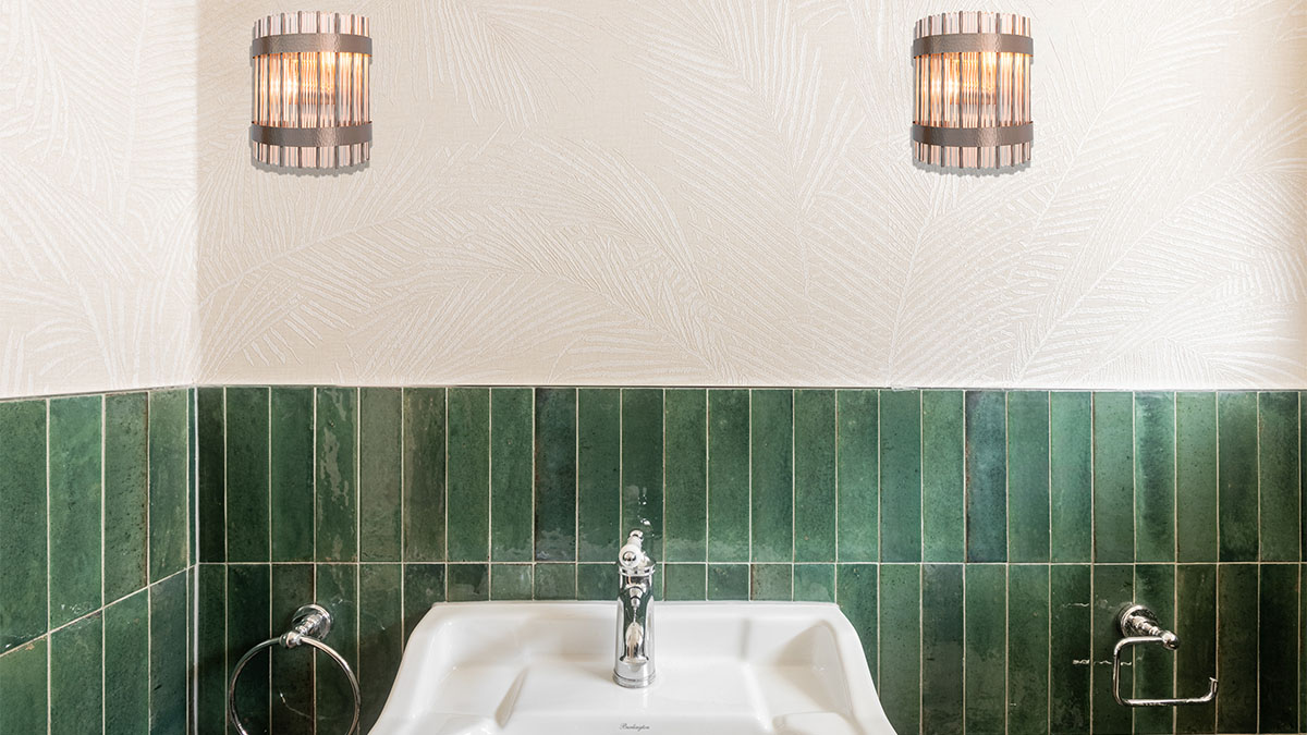 Stylish bathroom vanity area in a London flat with a white sink, chrome fixtures, dark green vertical tiles, and patterned wallpaper above, reflecting a high-end renovation