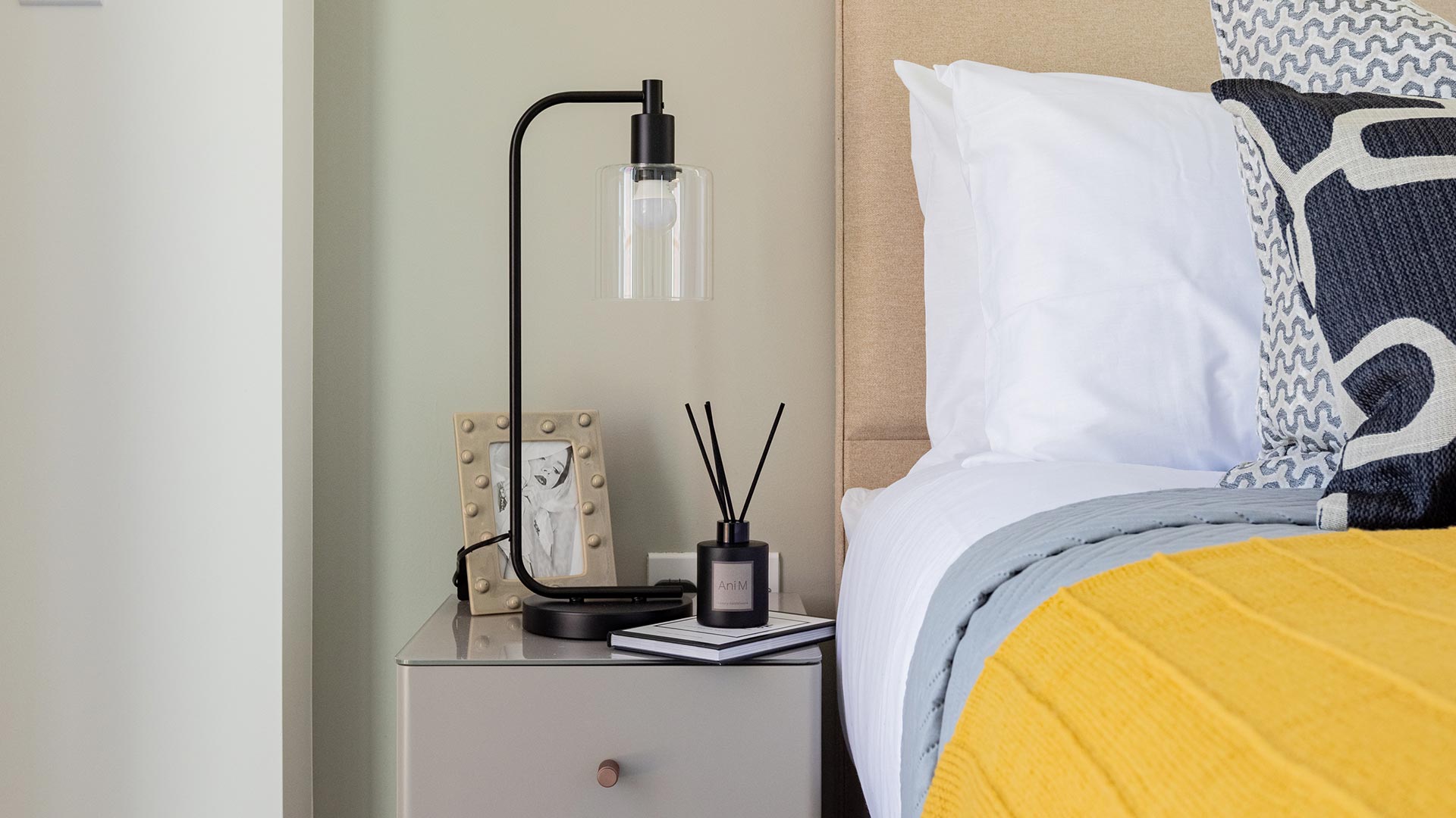 Close-up of a bedside table with a modern lamp, framed picture, and a diffuser, next to a bed with a yellow throw in Meridian One
