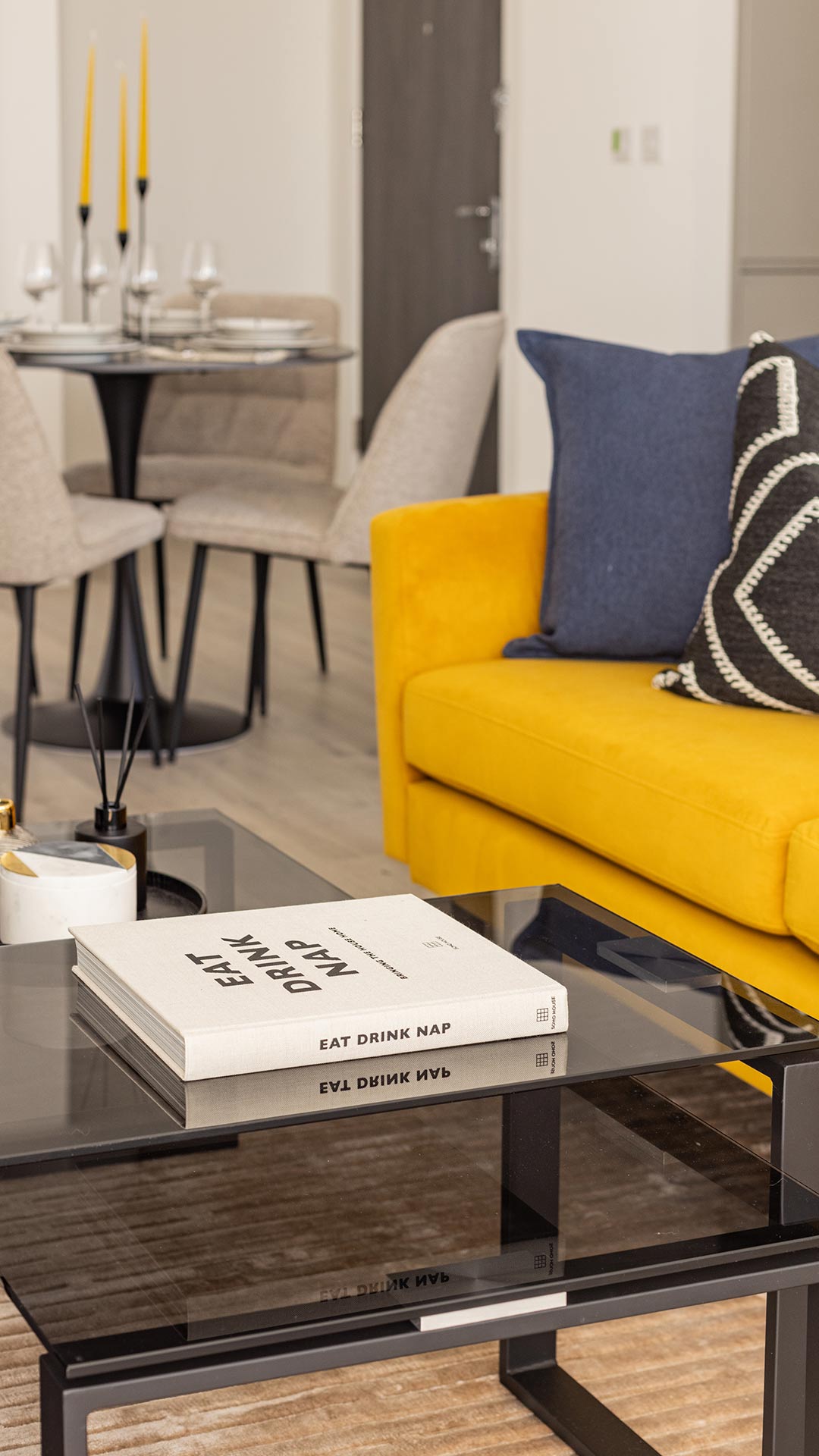 Modern glass coffee table with a book and decor items, reflecting a yellow sofa and dining area in a Meridian One London flat