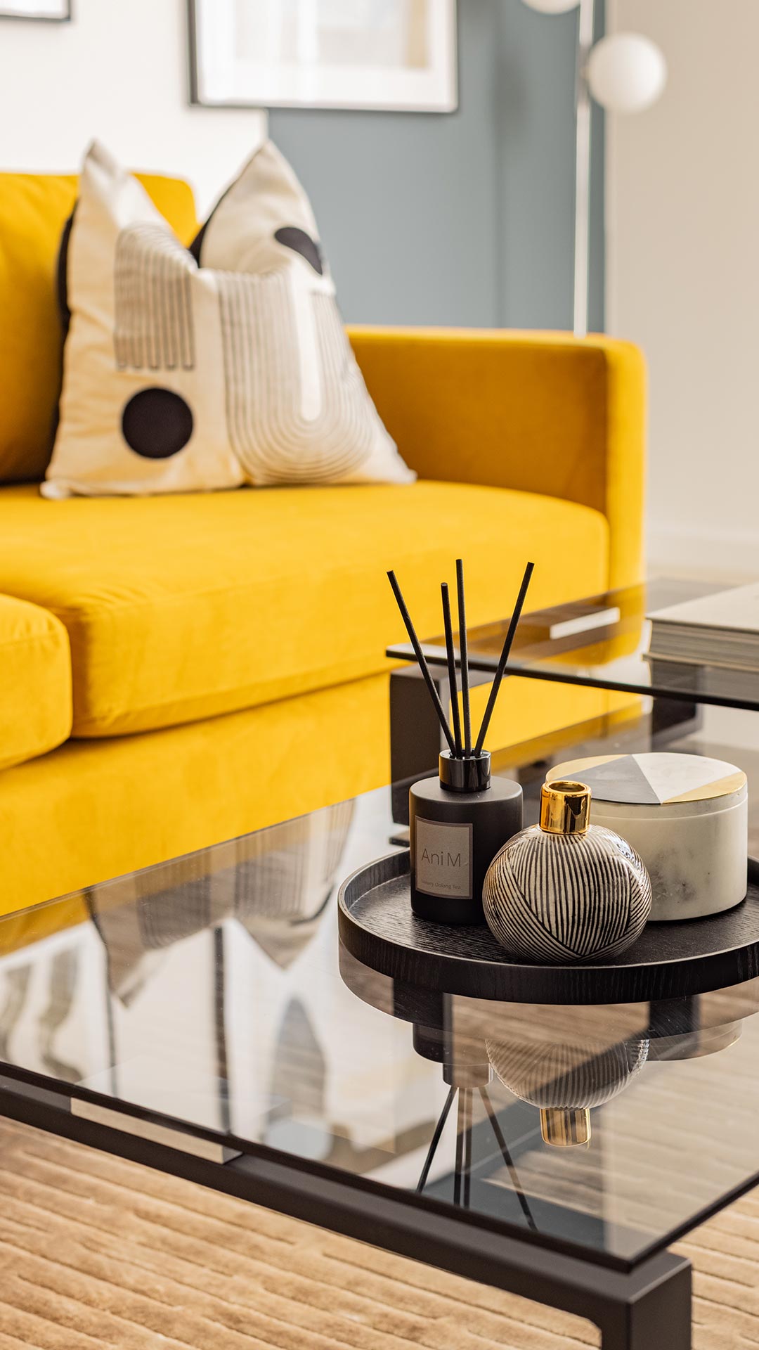 Close-up of a glass coffee table with a diffuser and decorative objects, in front of a yellow sofa in a Meridian One London flat