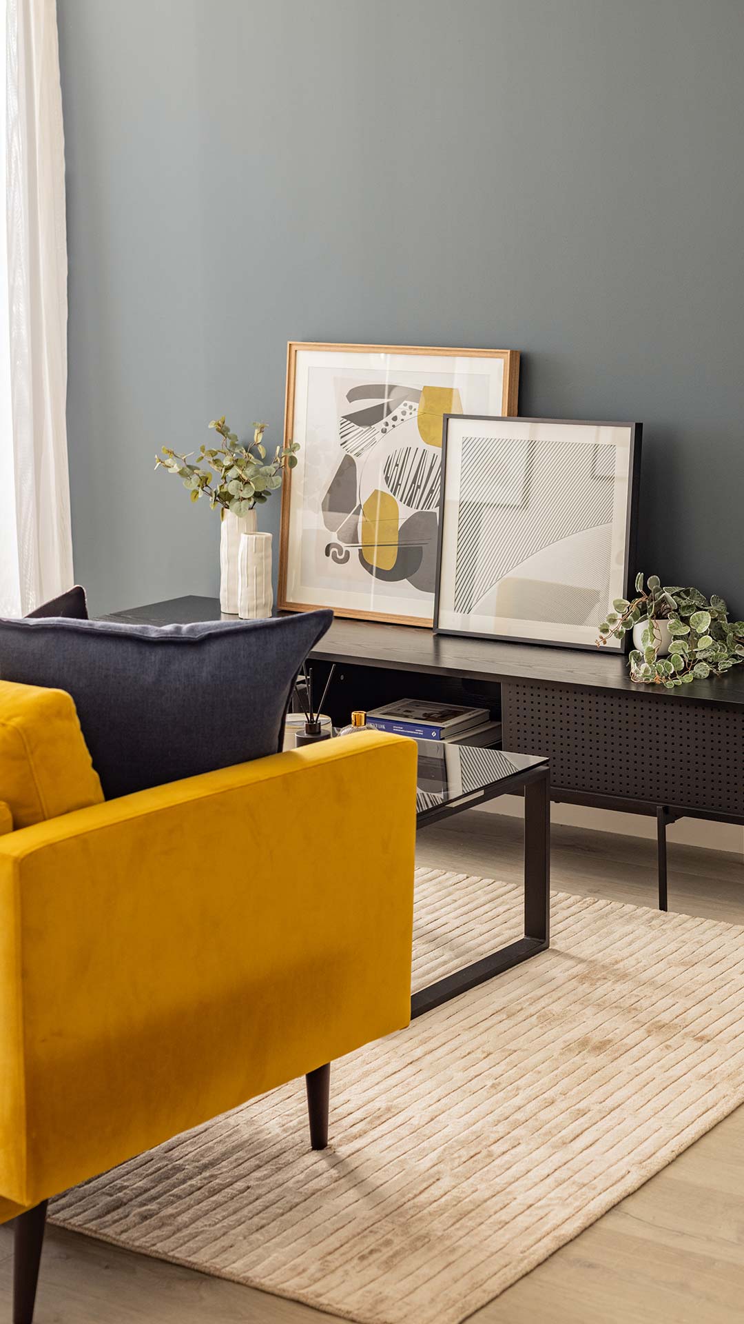 Black media unit with framed abstract art and plants, next to a yellow sofa, in a professionally decorated Meridian One living room