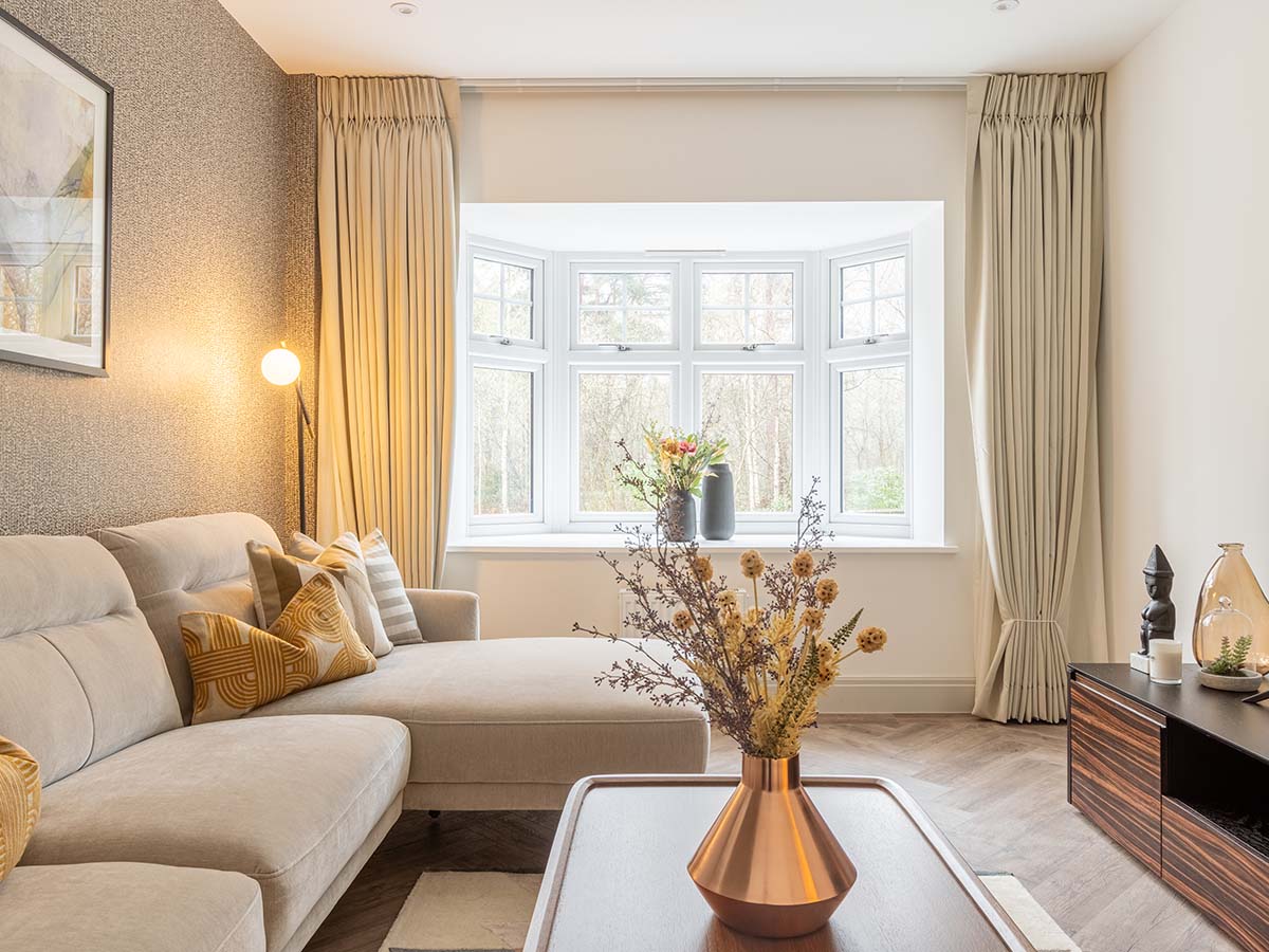 A cosy living room with a textured feature wall, a neutral L-shaped sofa with yellow patterned cushions, and a large bay window