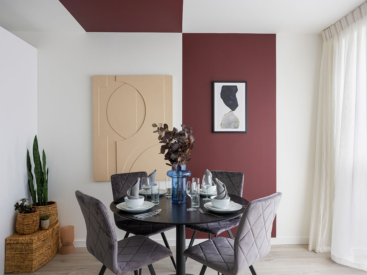 A modern dining area at Cargo House featuring a bold, colour-blocked burgundy feature wall, a black dining table, and quilted grey velvet chairs