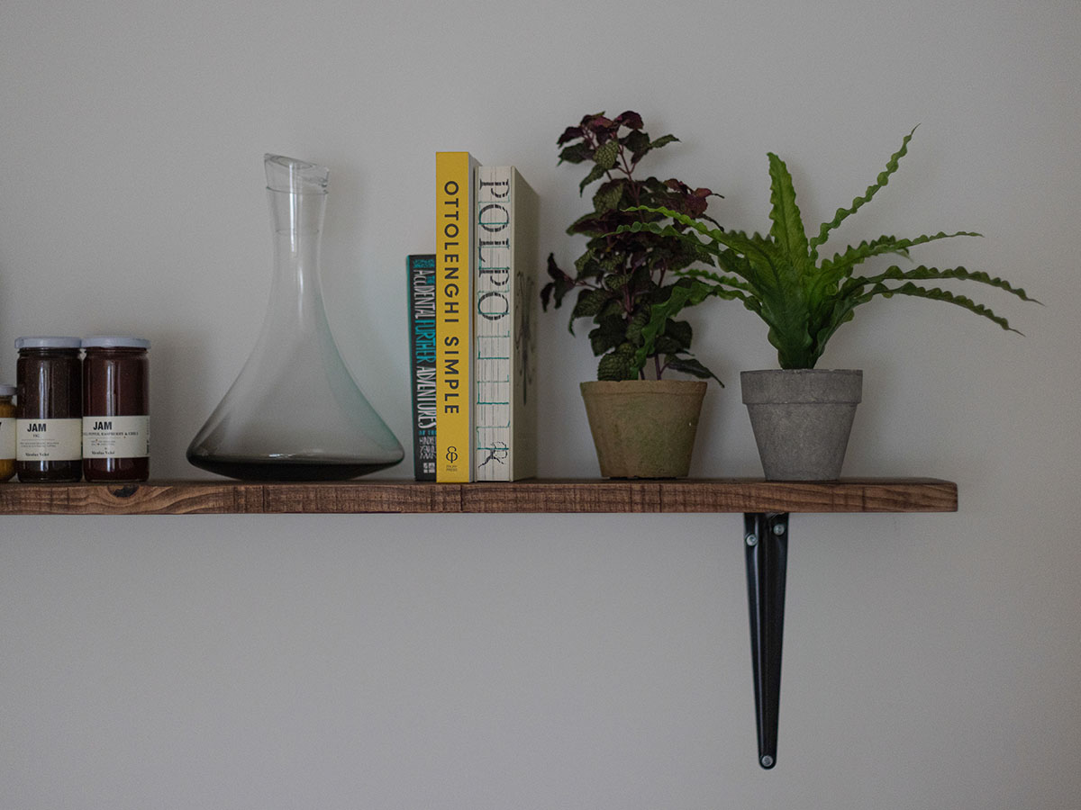 A close-up of a rustic wooden shelf styled with Ottolenghi cookbooks, a glass decanter, and small potted house plants