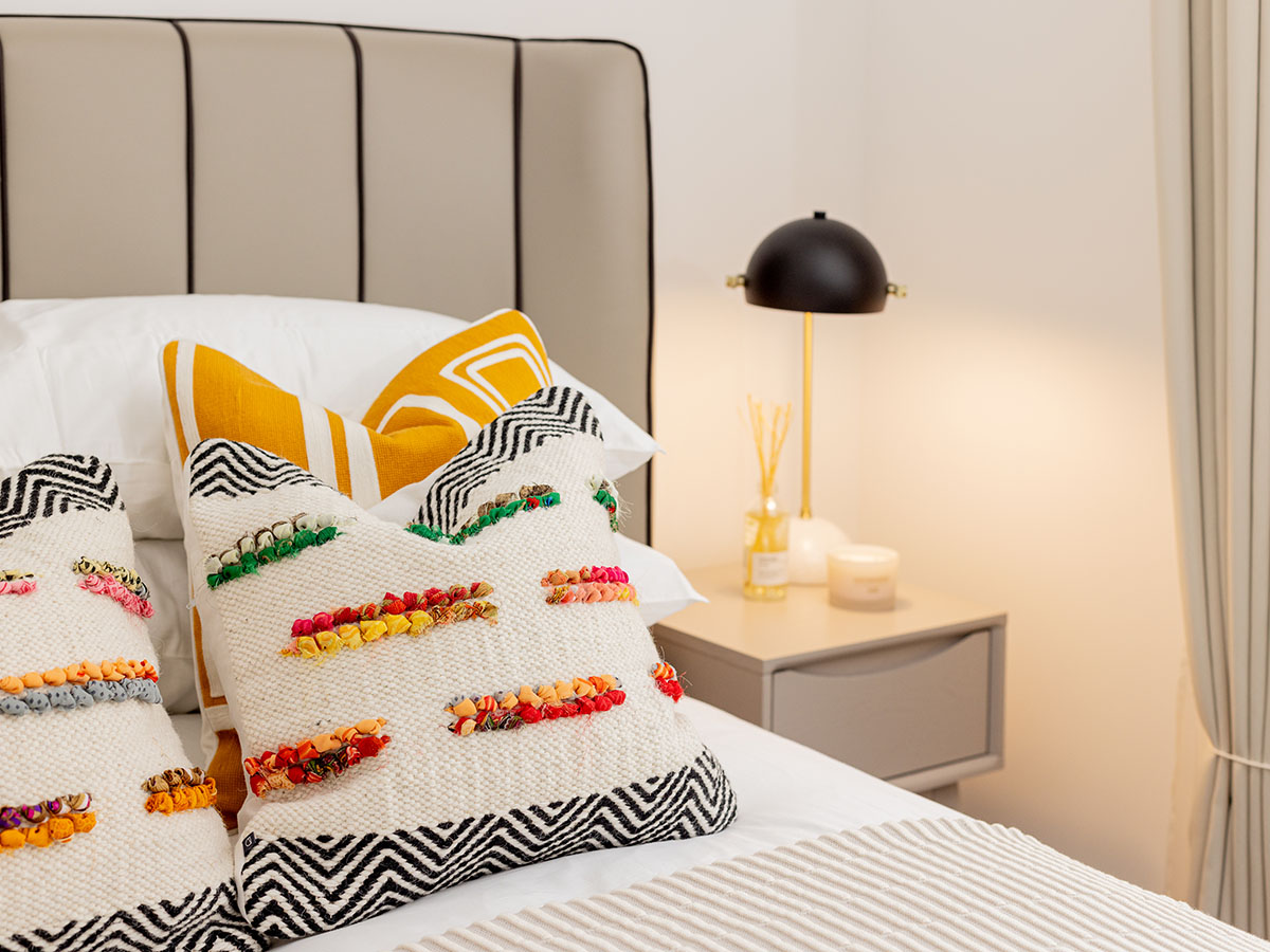 A close-up of a styled bed, showcasing highly textured woven cushions with colourful tufts against a yellow geometric pillow