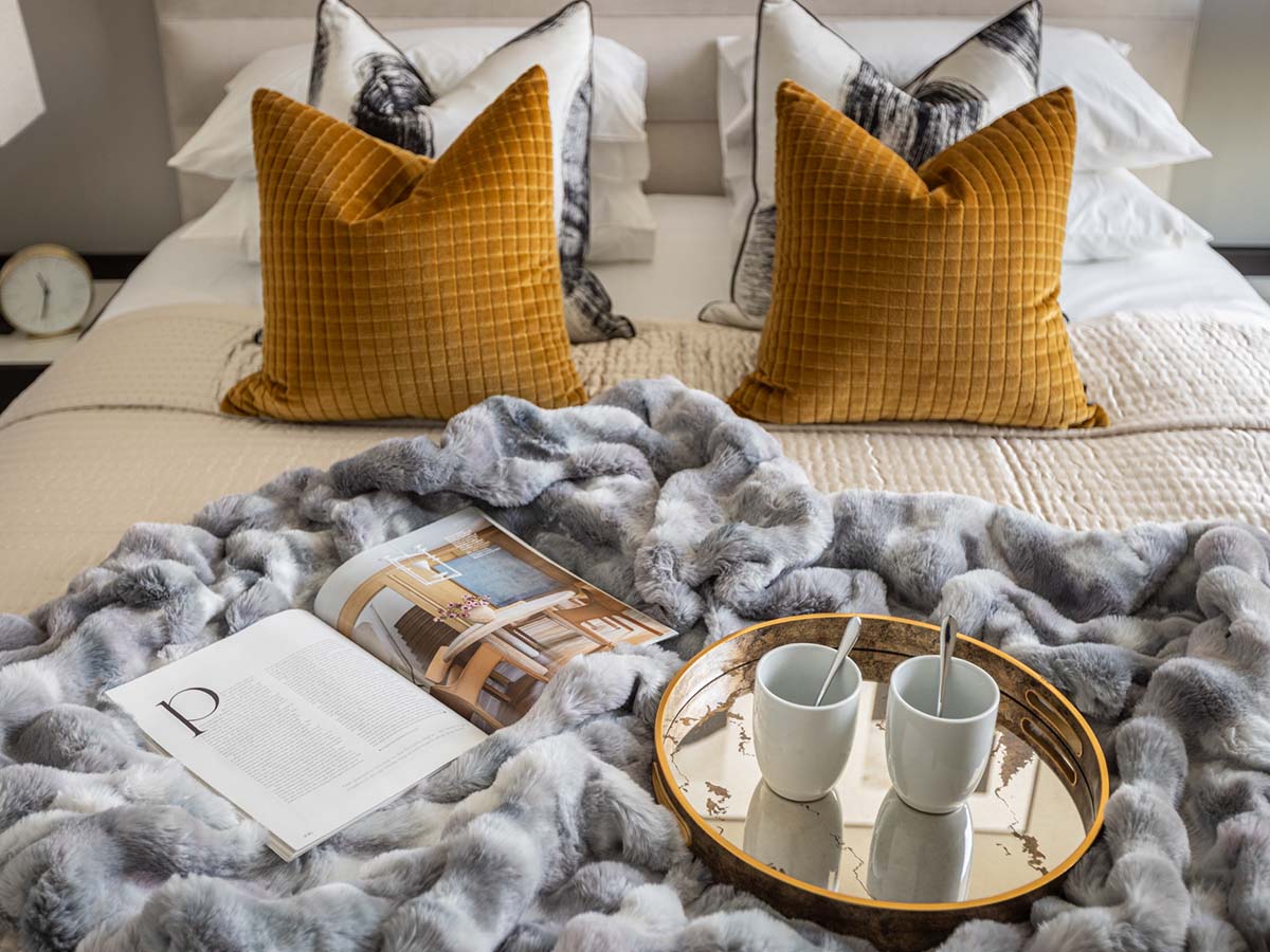 A luxuriously styled bed designed for homeowners, featuring mustard velvet cushions, a grey faux fur throw, and a breakfast tray for ultimate comfort
