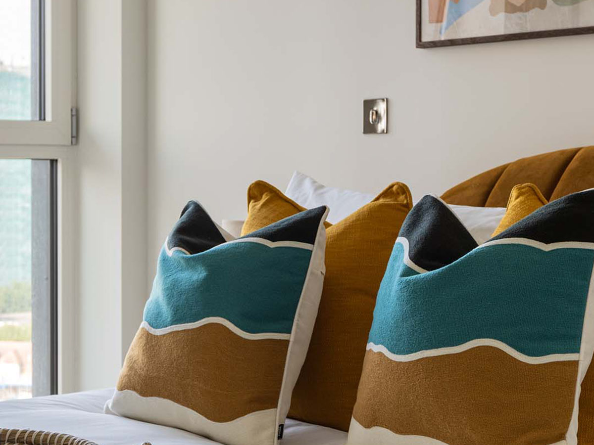 A close-up of a bed at White City Living, styled with bold, colour-blocked cushions in teal and brown on a channelled velvet headboard.