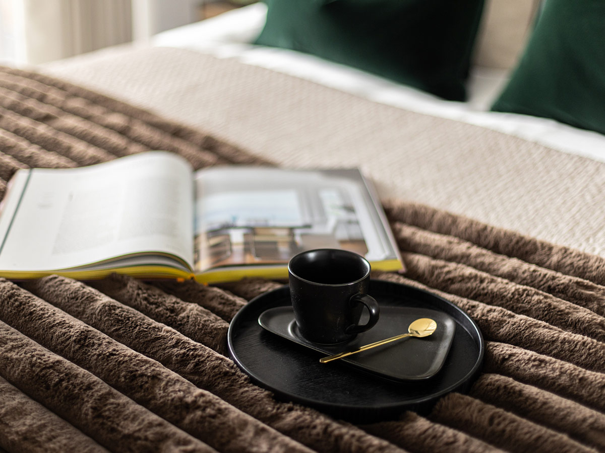 A cosy lifestyle detail shot in a bedroom at The Auria, showing a cup of coffee and an open book on a soft, textured brown throw.