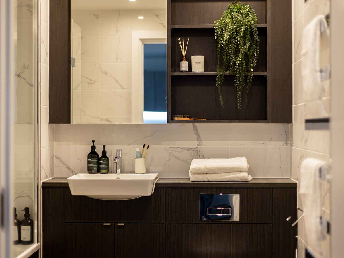 A modern bathroom at The Auria featuring a dark wood vanity, white marble-effect tiles, and a stylishly arranged open shelf with a trailing plant.