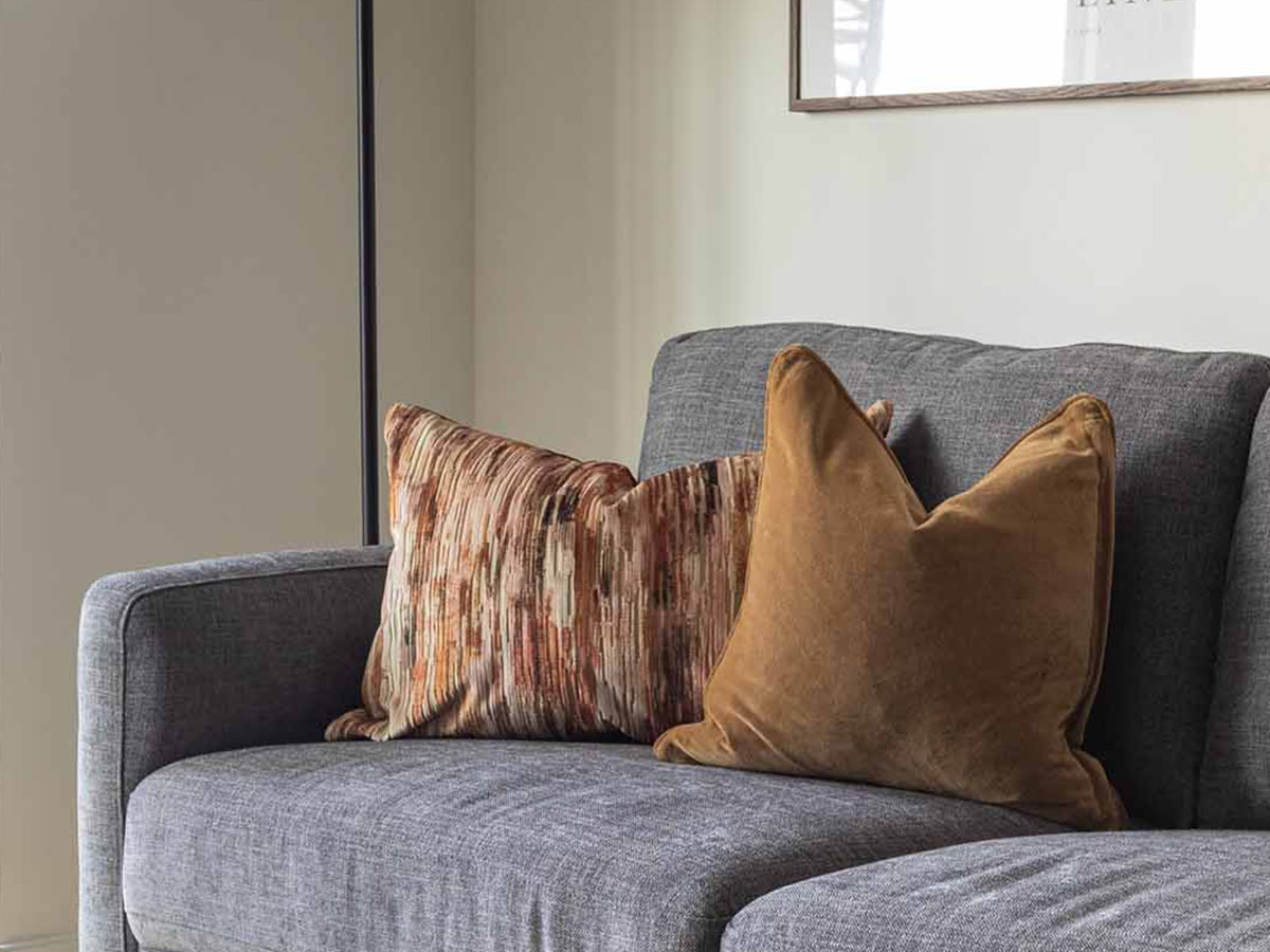 A close-up of a stylishly dressed grey sofa at White City Living, featuring a mix of golden-brown velvet and colourful abstract patterned cushions.
