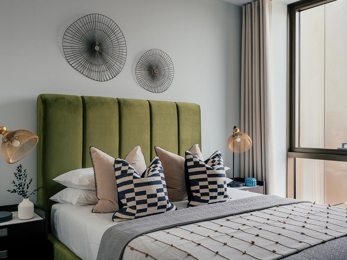 An aspirational show home bedroom designed for property developers, featuring a bold olive green channel-tufted headboard, wire wall art, and gold mesh lamps