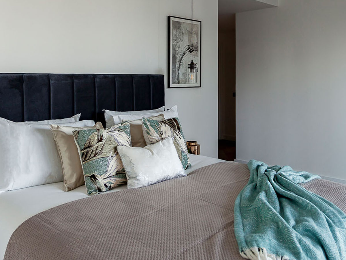 A sophisticated bedroom at Merina House, London Dock, featuring a tall, dark velvet channel-tufted headboard and botanical print cushions.
