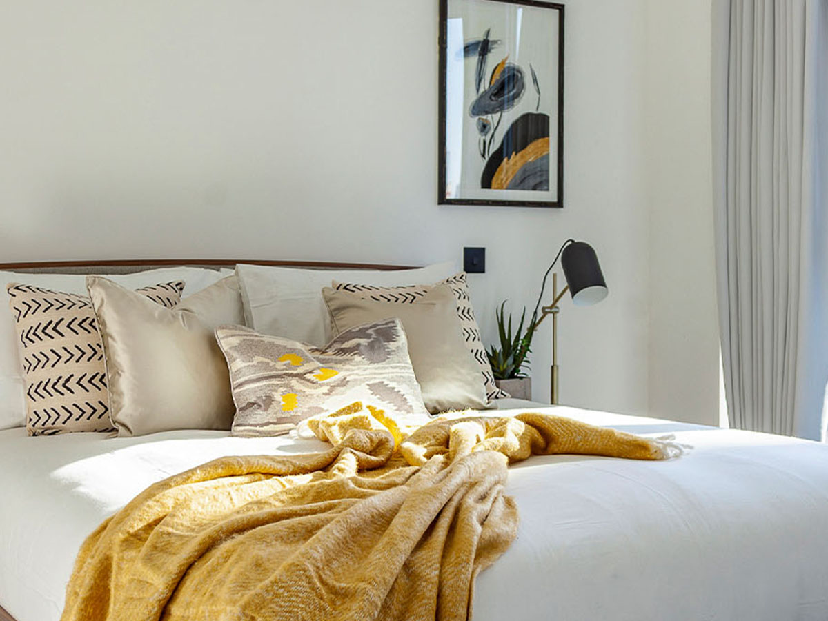 A bright, Scandi-style bedroom at Merina House, London Dock, styled with a mustard yellow throw and a mix of patterned cushions.