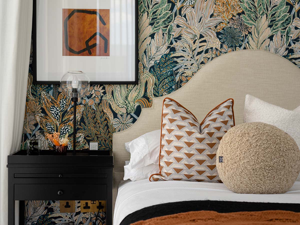 A maximalist bedroom design featuring a bold, colourful botanical wallpaper, a curved headboard, and a mix of textured and patterned cushions