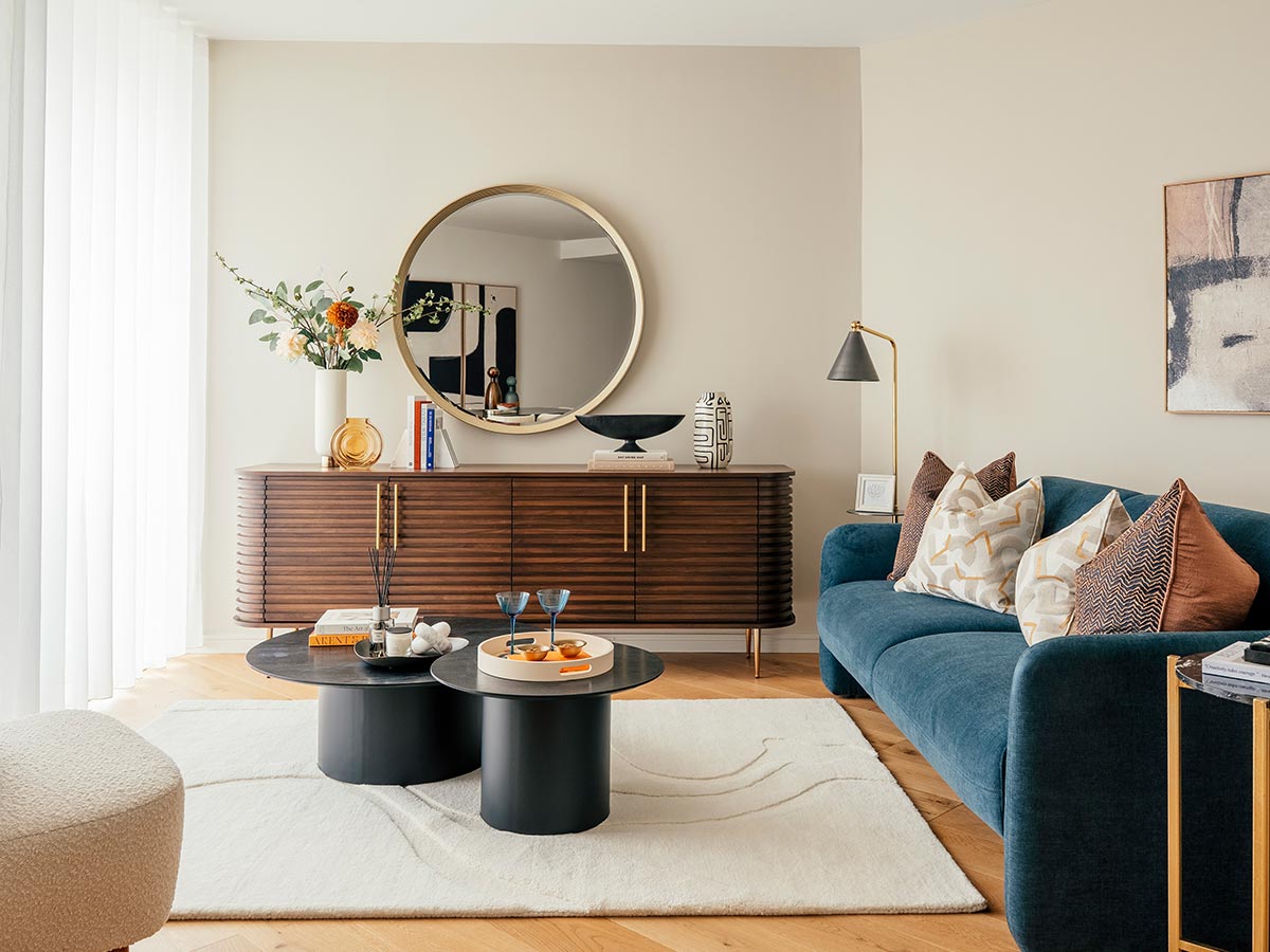 A professionally styled living room designed to help estate agents market properties, featuring an elegant blue sofa, a slatted wood console, and a large round mirror