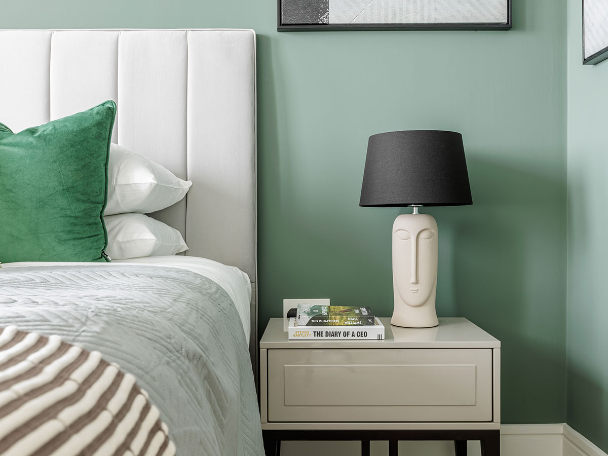 A serene bedroom at One Bishopgate with a sage green feature wall, a white channel-tufted headboard, and a modern sculptural face lamp