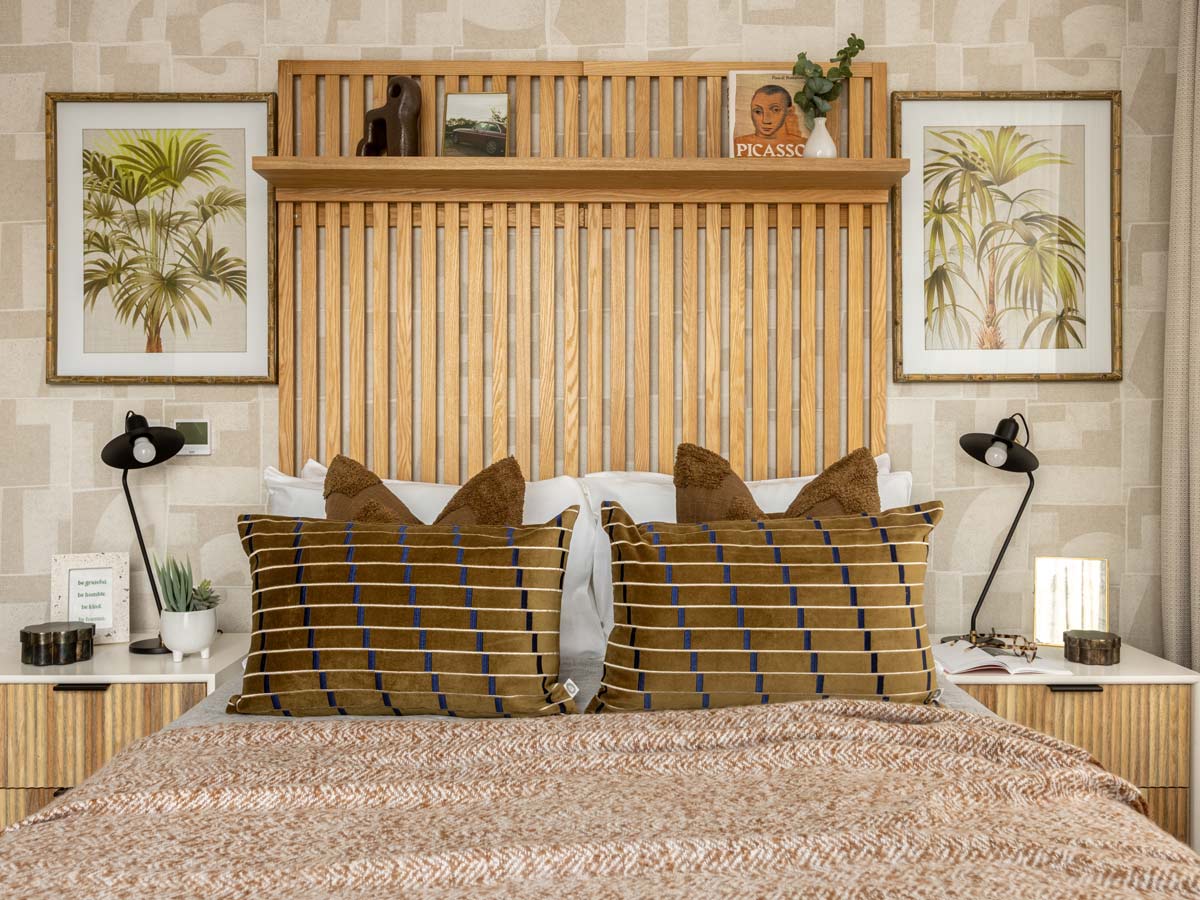 A stylish show home bedroom featuring a large slatted oak headboard with a display shelf, geometric wallpaper, and framed palm tree art