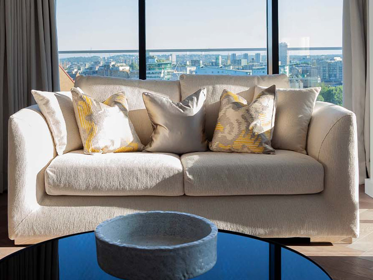 A comfortable cream sofa with yellow patterned cushions at Merina House, London Dock, set against a large window with panoramic London city views.