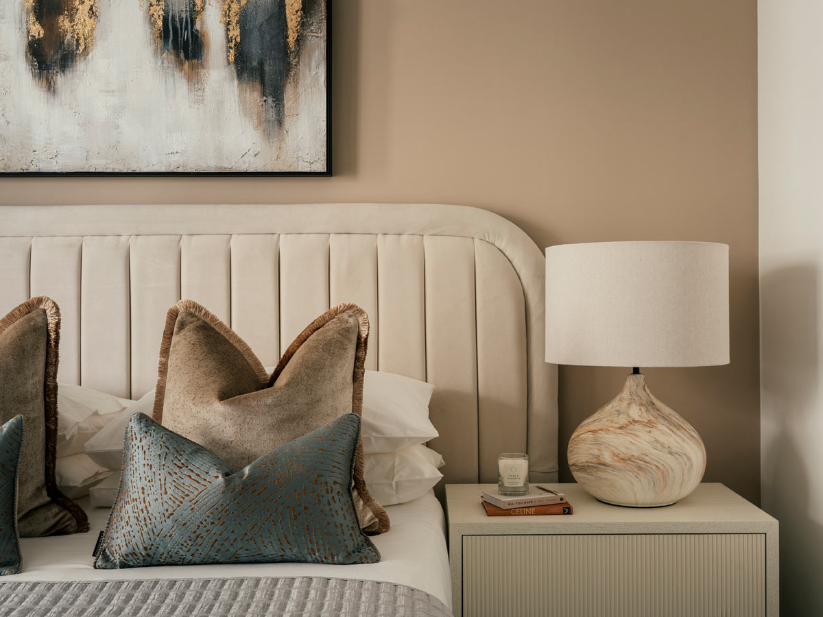 A close-up of a styled bed at the Riverside project, with a curved cream channel-tufted headboard, textured cushions, and a modern ceramic lamp on the bedside table