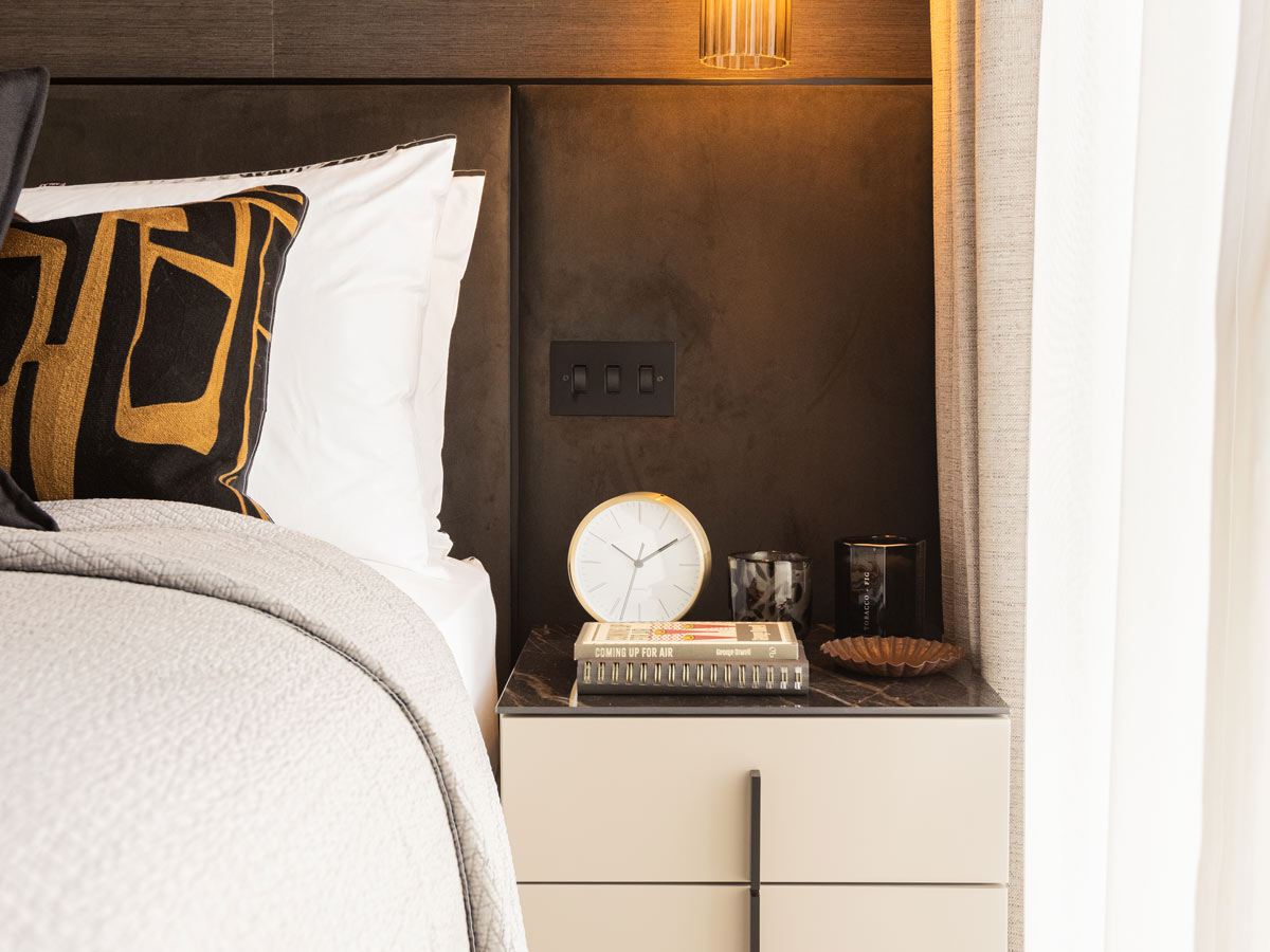 A close-up of a luxury bedroom at the Aspen project, showing a marble-topped bedside table styled with a clock, books, and a designer cushion on the bed