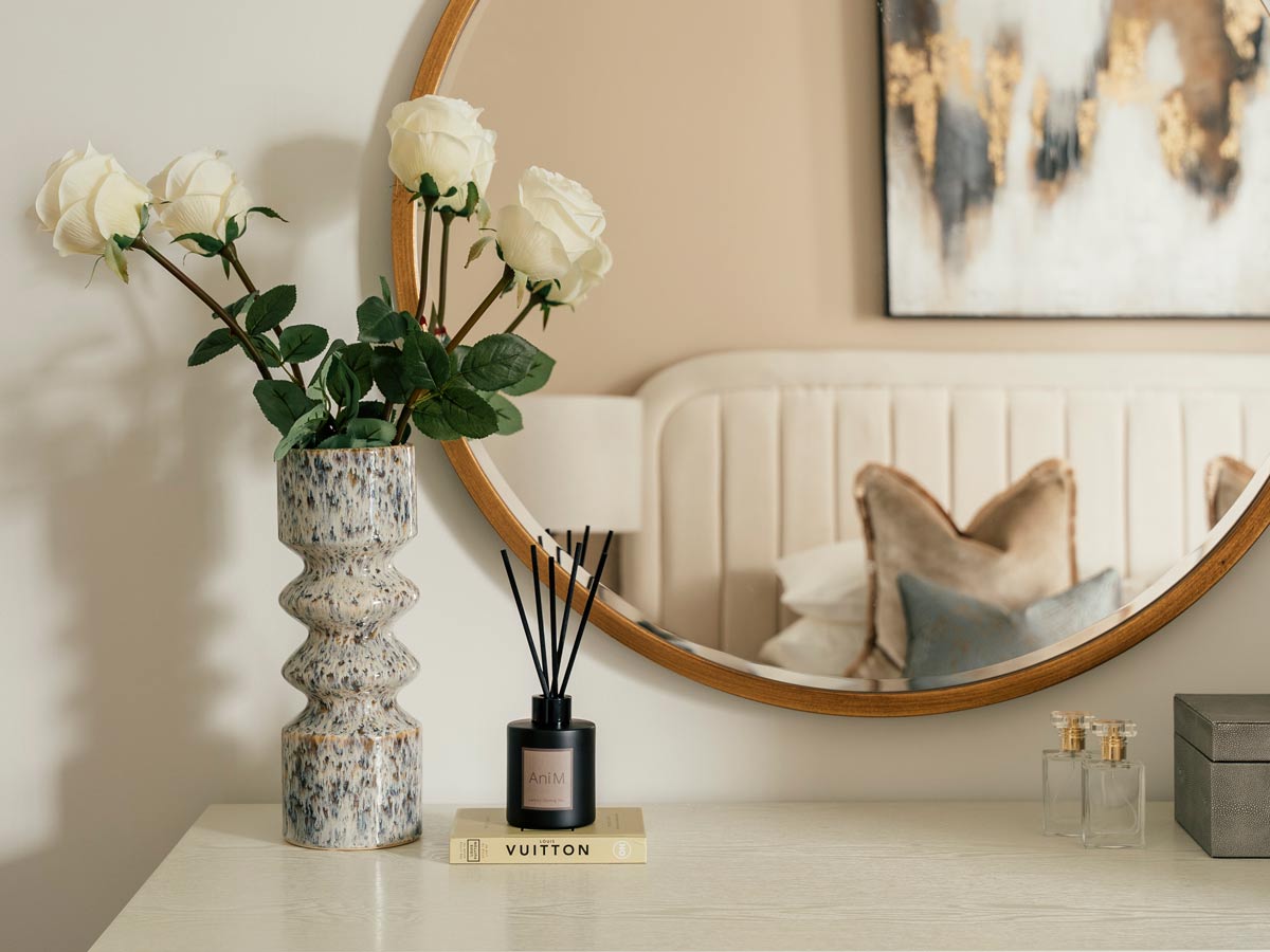 A detailed shot of a white bedroom dresser at the Riverside project, styled with a ceramic vase of white roses and a reed diffuser, with a large round mirror reflecting the bed