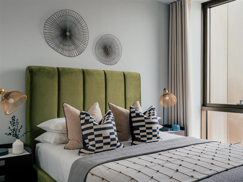 A stylish modern bedroom at the Riverside project, featuring a tall, olive green velvet headboard, geometric cushions, wire wall art, and contemporary gold mesh bedside lamps