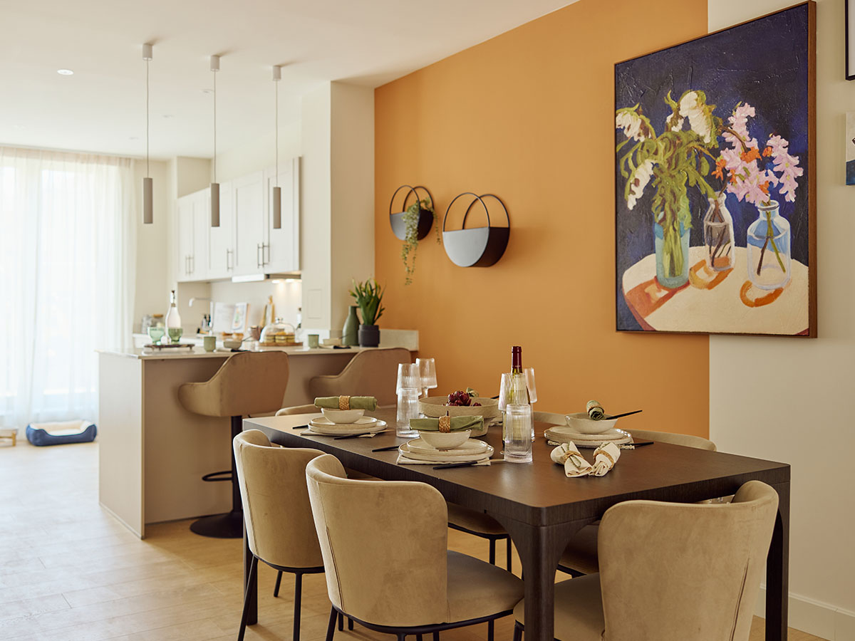 An open-plan dining area at New Fountainbridge, featuring a set dining table, a bold orange accent wall with a large floral painting, and a view to the kitchen island