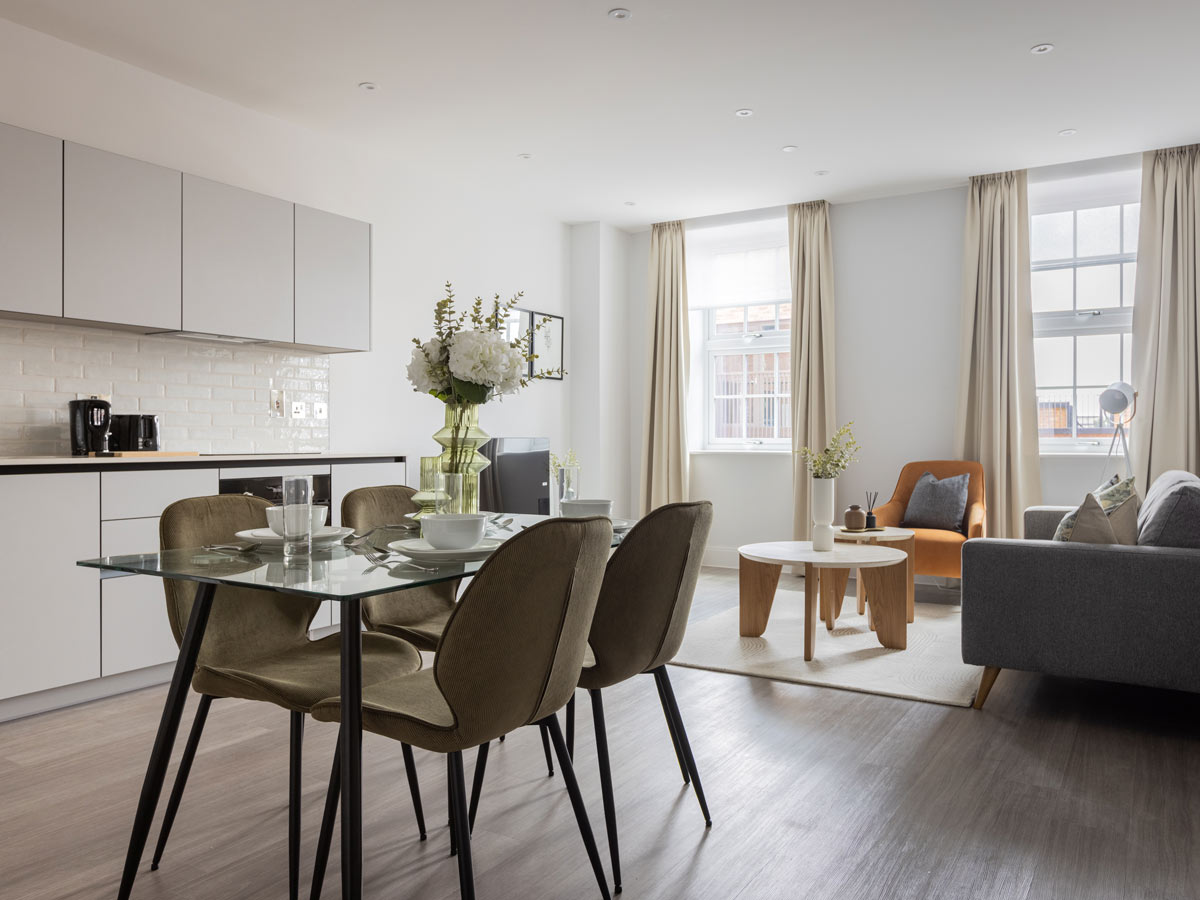 A bright, open-plan apartment at The Factory, Horlicks Quarter, featuring a modern white kitchen, a glass dining table, and a cosy living area