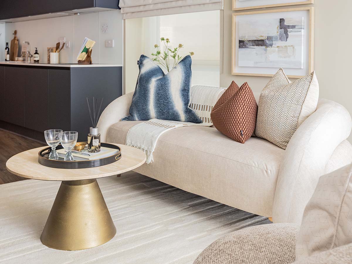 A modern living area featuring a light-colored, curved sofa accented with blue tie-dye and terracotta pillows, a central conical gold coffee table, and an open-plan dark kitchen in the background.