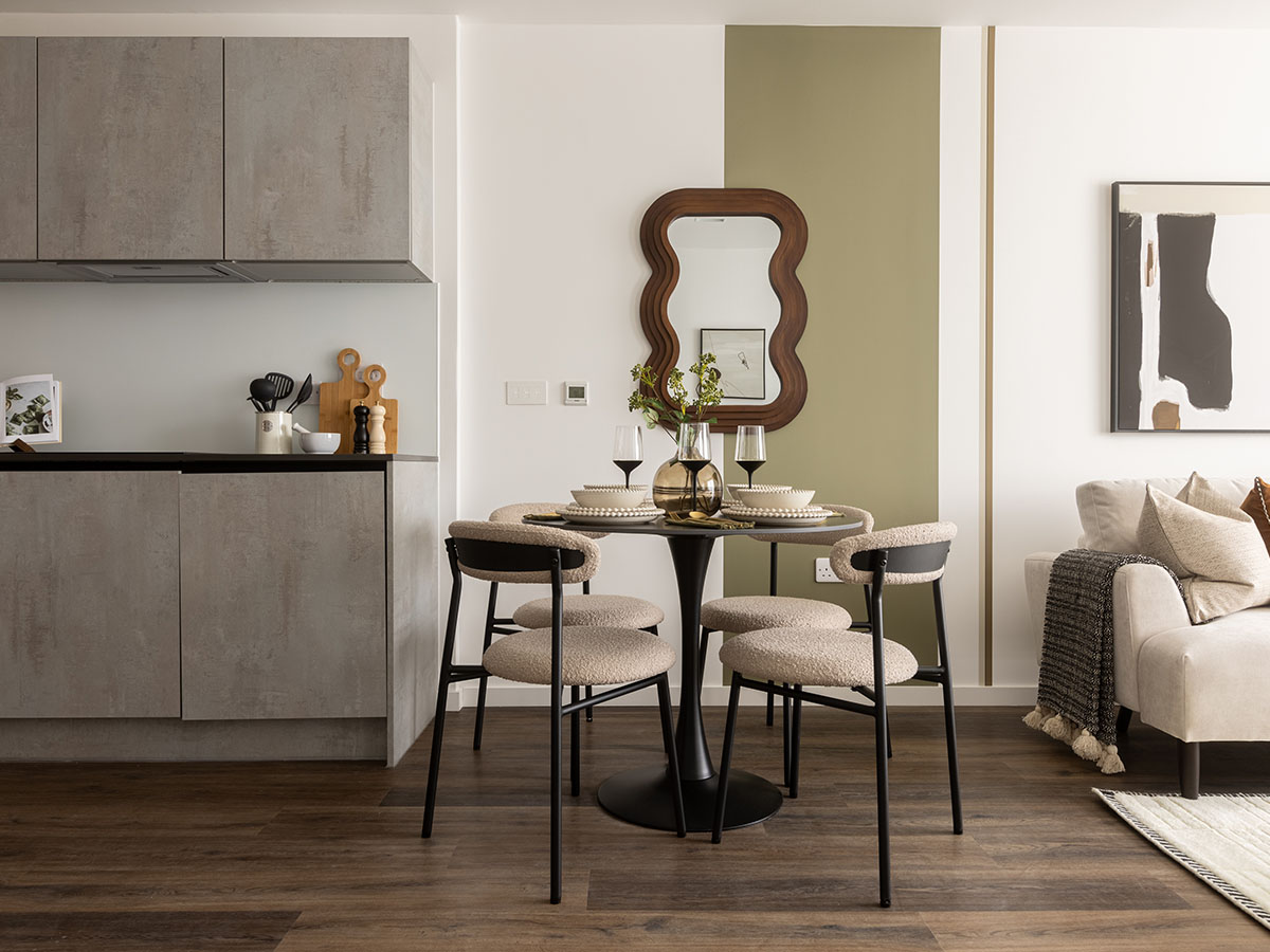 A chic open-plan kitchen and dining area at The Auria, featuring stone-effect grey cabinets, a round black dining table with beige bouclé chairs, and a vertical olive-green feature stripe on the wall