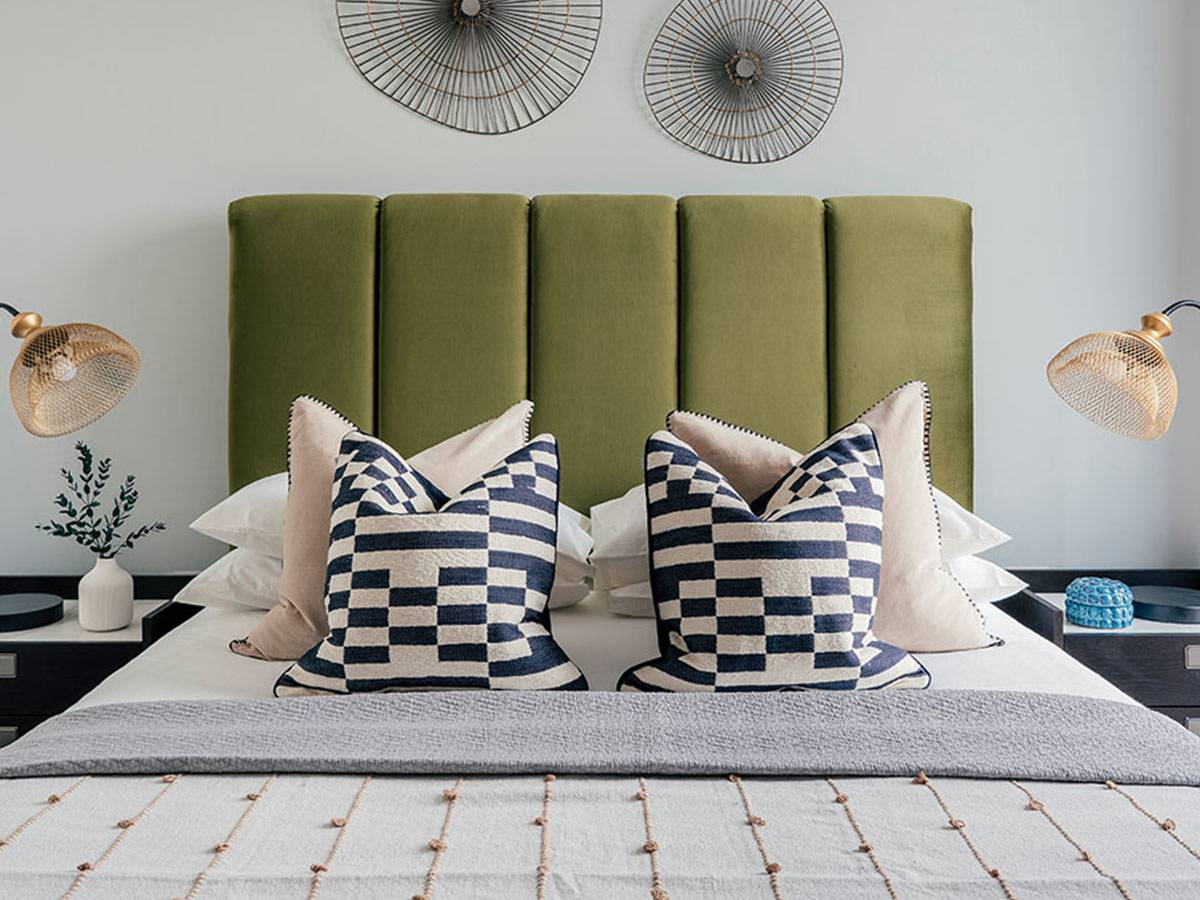 A symmetrical bedroom at 804 Riverside featuring an olive green velvet headboard, geometric cushions, gold mesh lamps, and wire wall art