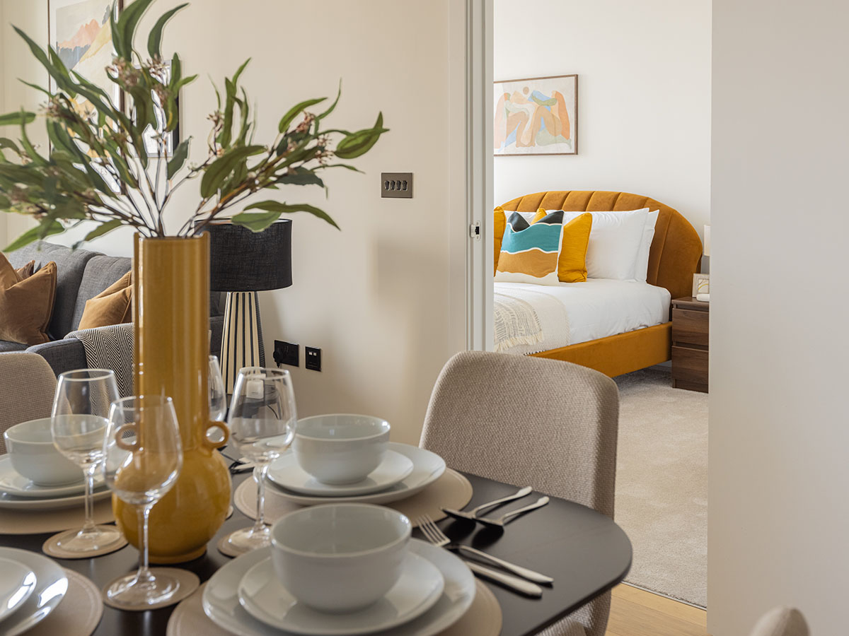 A dining table setting at White City with white bowls and a yellow vase, providing a view into a bedroom featuring a mustard yellow tufted headboard