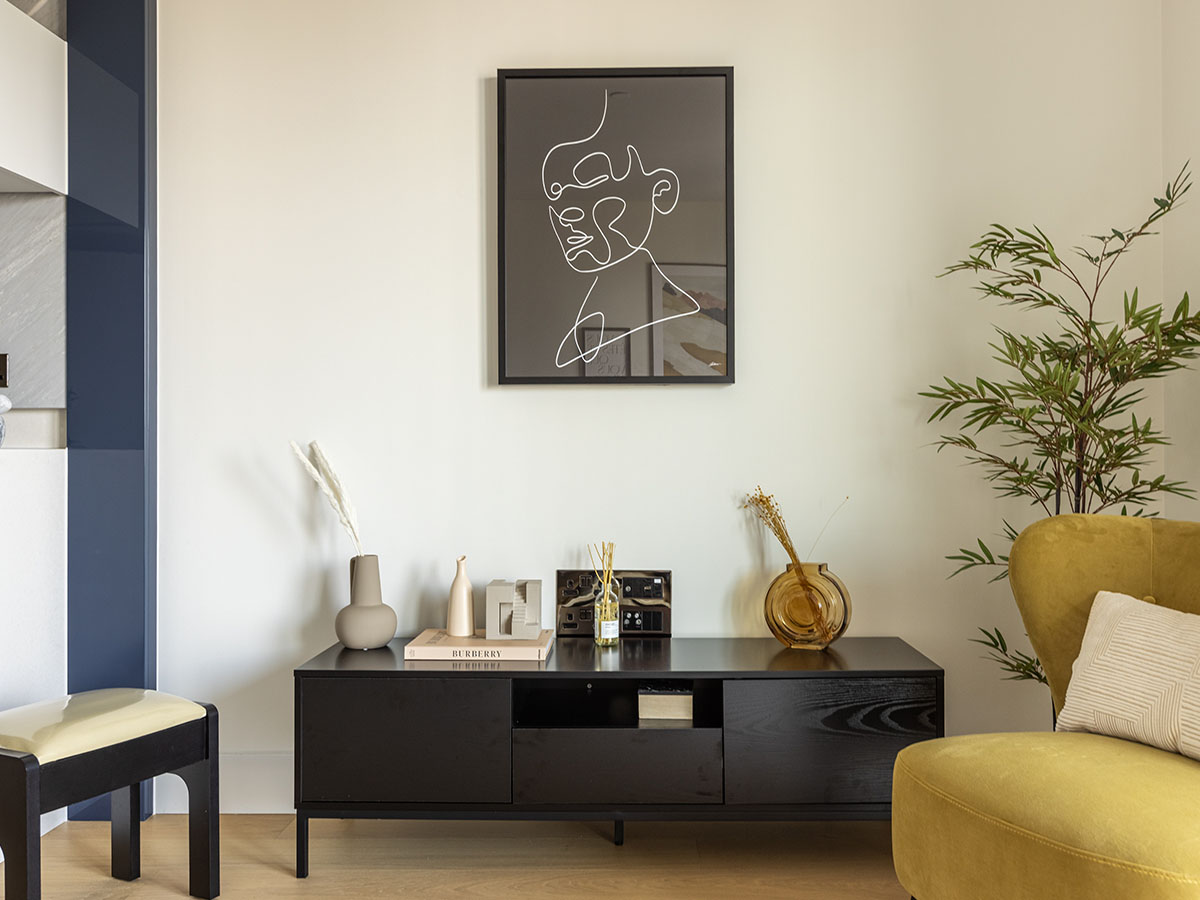 A minimalist media console setup at White City featuring a black wooden sideboard with decorative vases, a line-art portrait on the wall, and a yellow wingback chair