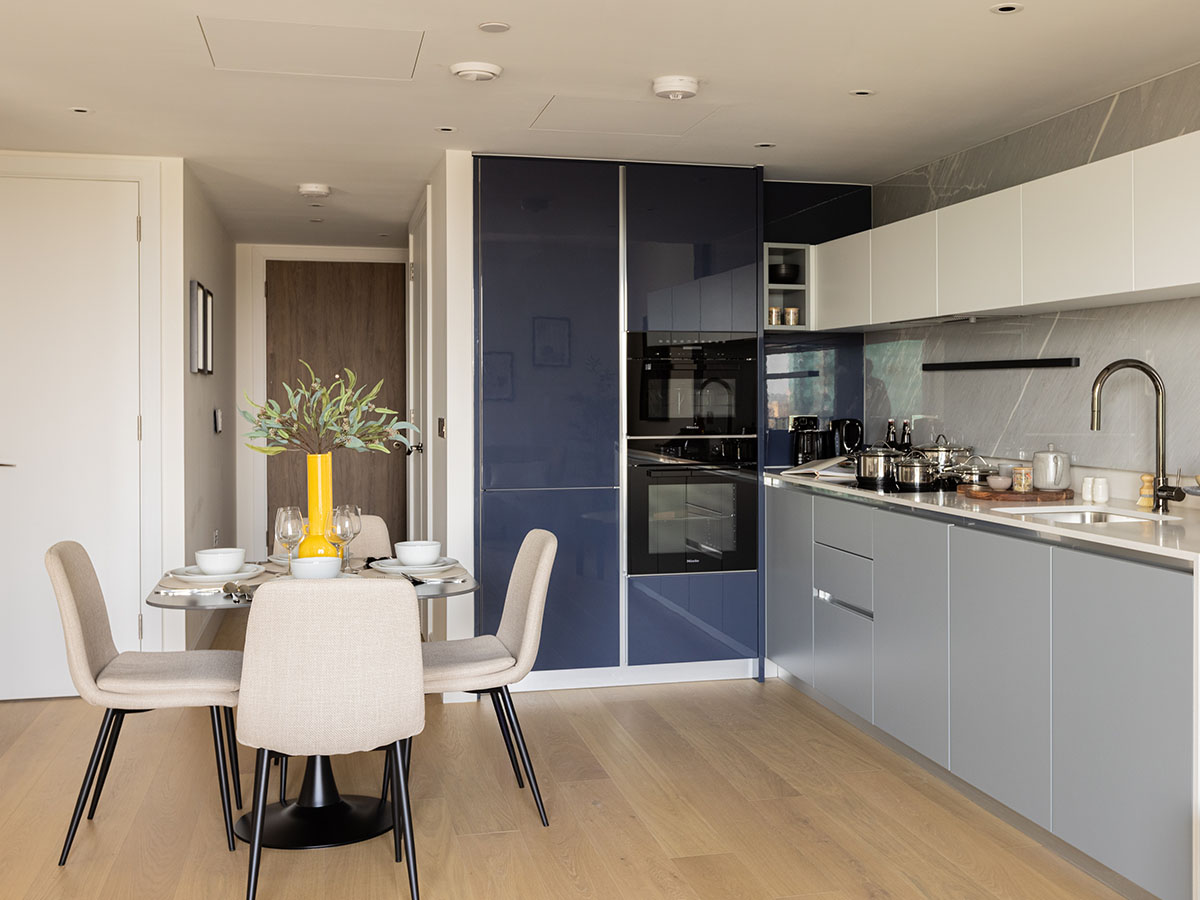 A sleek modern kitchen and dining area at White City featuring navy blue cabinetry, light grey lower cabinets, and a dining table with neutral chairs and a yellow vase