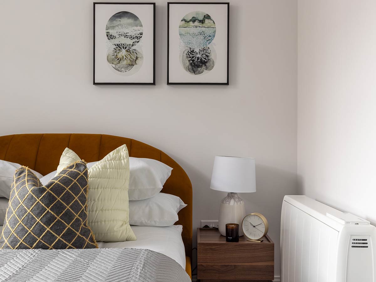 A master bedroom in the Horlicks Quarter, Slough, featuring an amber velvet scalloped headboard, geometric patterned cushions, a dark wood nightstand with a marble lamp, and abstract botanical artwork