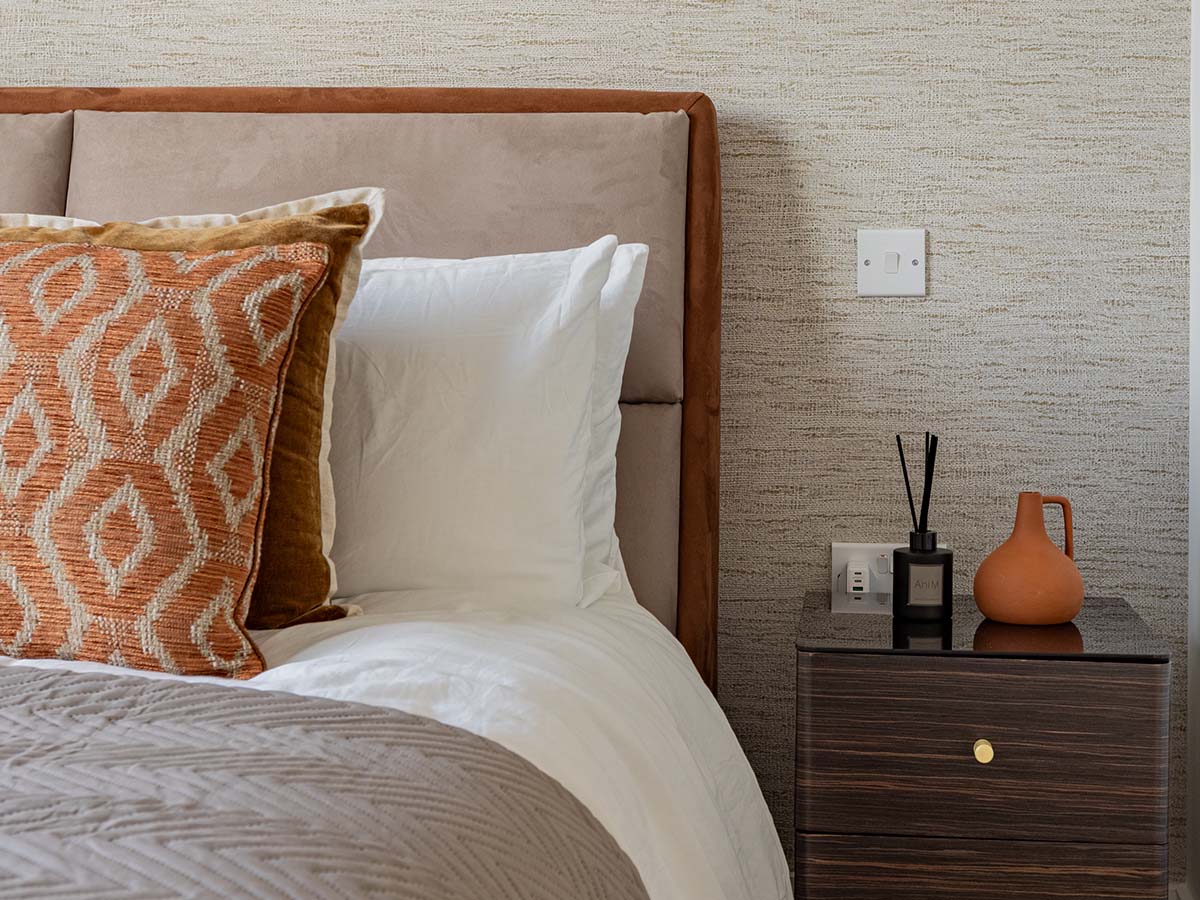 Close-up of a bespoke bedroom interior featuring a plush taupe headboard, white linens with orange geometric patterned cushions, and a dark wood bedside table with a designer reed diffuser and terracotta vase