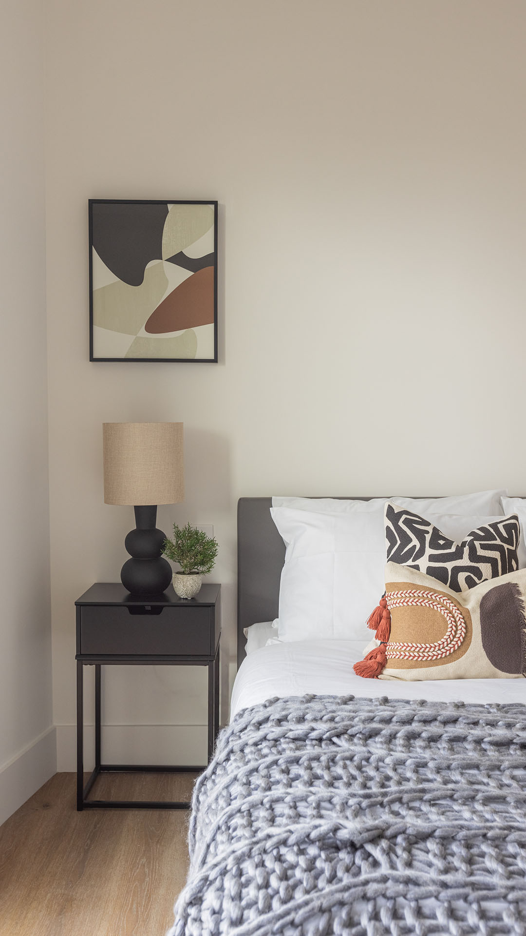 A minimalist bedroom corner featuring a matte black bedside table and sculptural lamp, topped with an earth-toned abstract framed print above a grey upholstered headboard.