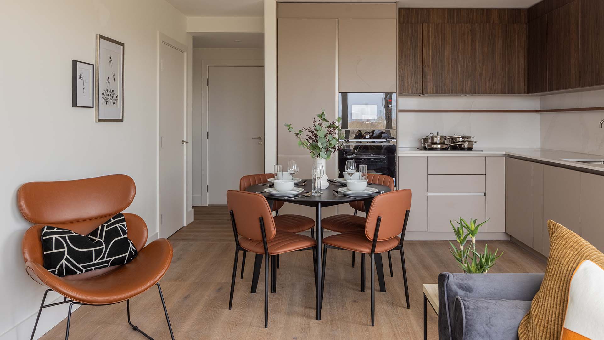 Wide-angle view of a professionally furnished apartment showing the kitchen, dining zone with tan leather seating, and a designer tan accent chair.