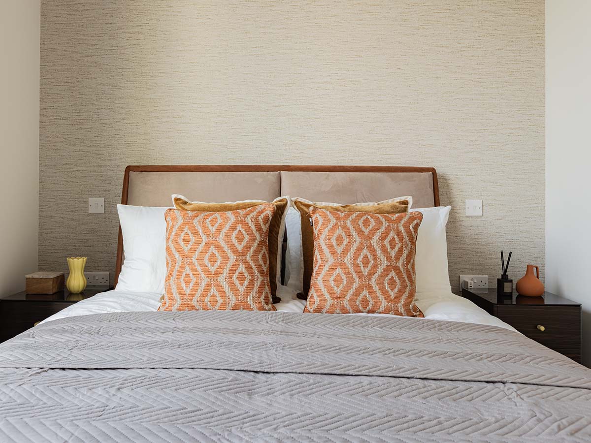 Symmetrical view of a luxury hotel-style bedroom featuring a soft-padded headboard, vibrant orange cushions, and matching dark wood nightstands against a textured feature wall