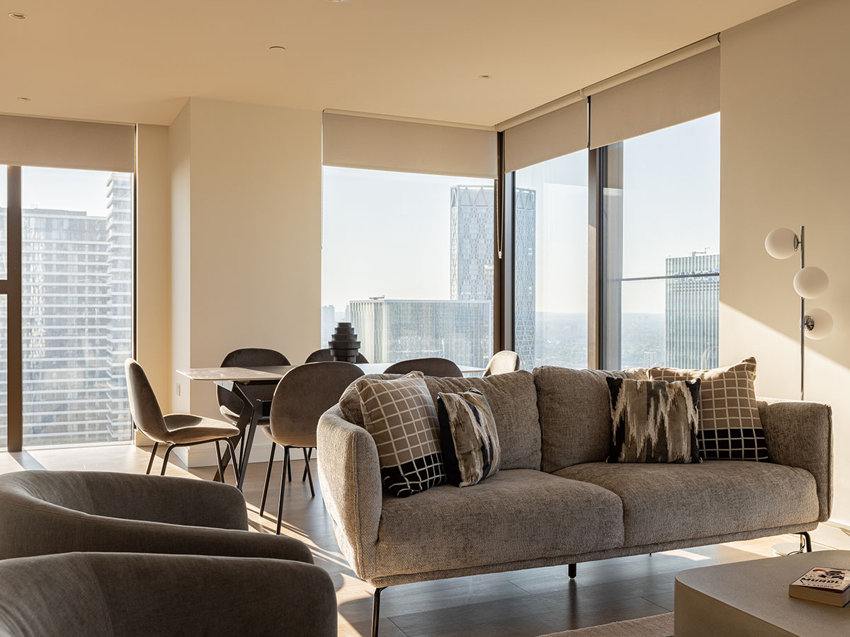 A luxury modern living room designed by InStyle Direct featuring a textured grey sofa with geometric cushions, a marble-top dining table with velvet chairs, and large windows overlooking the Canary Wharf skyline in London