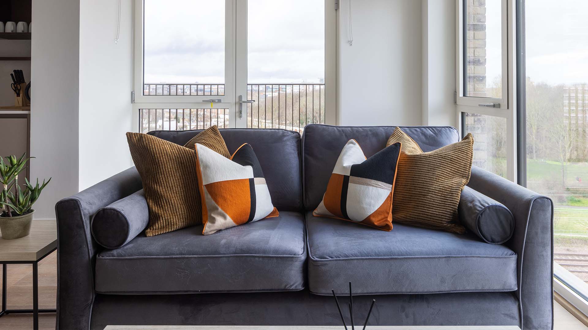 Front-facing view of a plush grey velvet sofa adorned with ochre and geometric patterned cushions, set against expansive windows overlooking a London urban landscape.