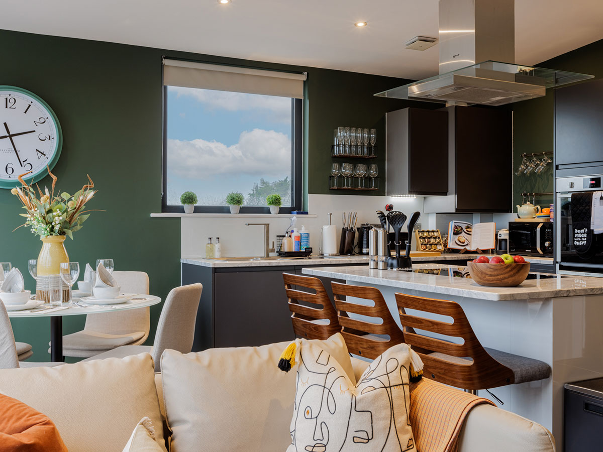 A luxury open-plan kitchen and dining area styled by InStyle Direct featuring deep green walls, a marble-top kitchen island with wooden bar stools, a round dining table with neutral chairs, and a large teal wall clock in Fulham