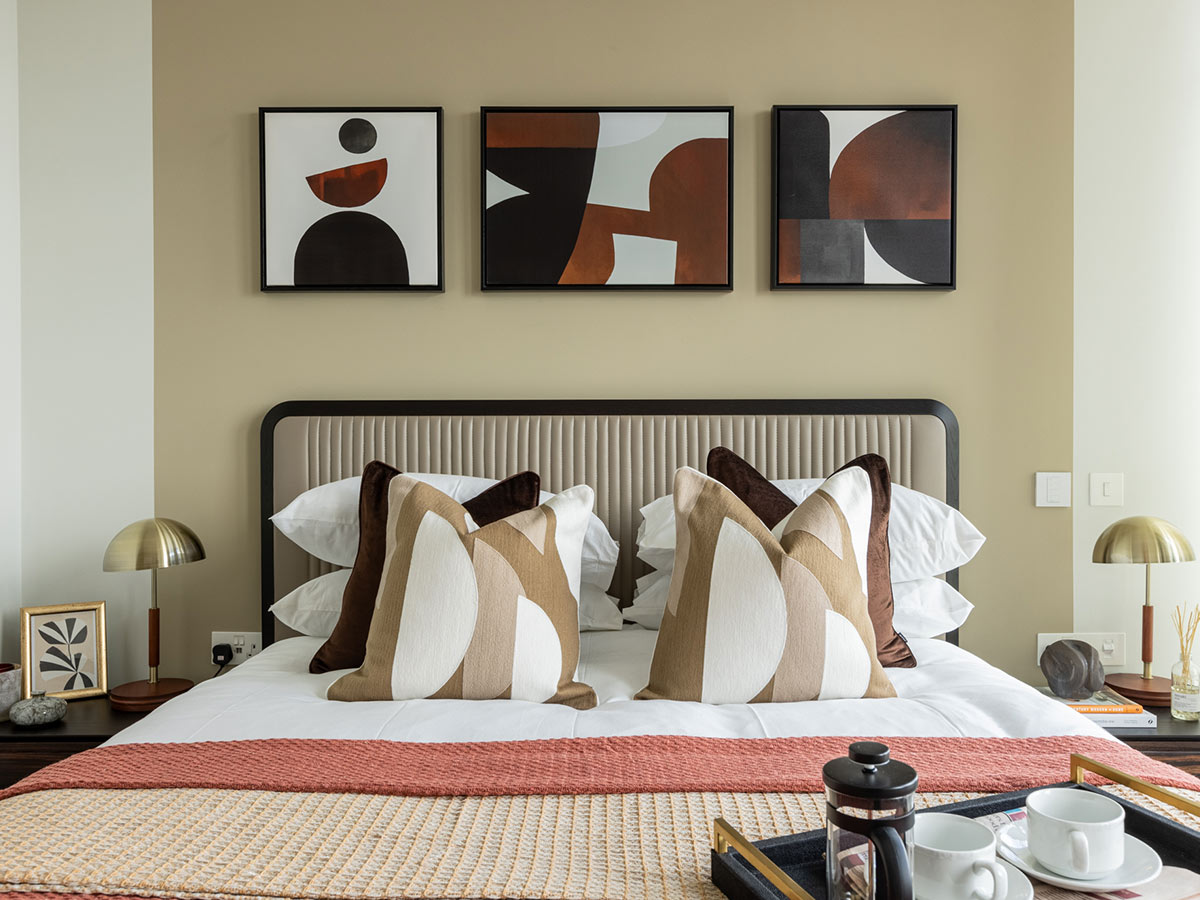 A bespoke bedroom interior styled by InStyle Direct featuring a neutral ribbed headboard, symmetrical brass bedside lamps, three framed abstract geometric art pieces in earthy tones, and a breakfast tray set on a white duvet in Shoreditch
