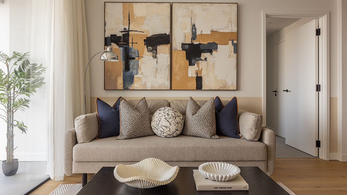 A contemporary living room at Koa House styled by InStyle Direct featuring a neutral sofa with navy and patterned cushions, a large two-panel abstract artwork in earth tones, and a sleek black coffee table with decorative fluted bowls