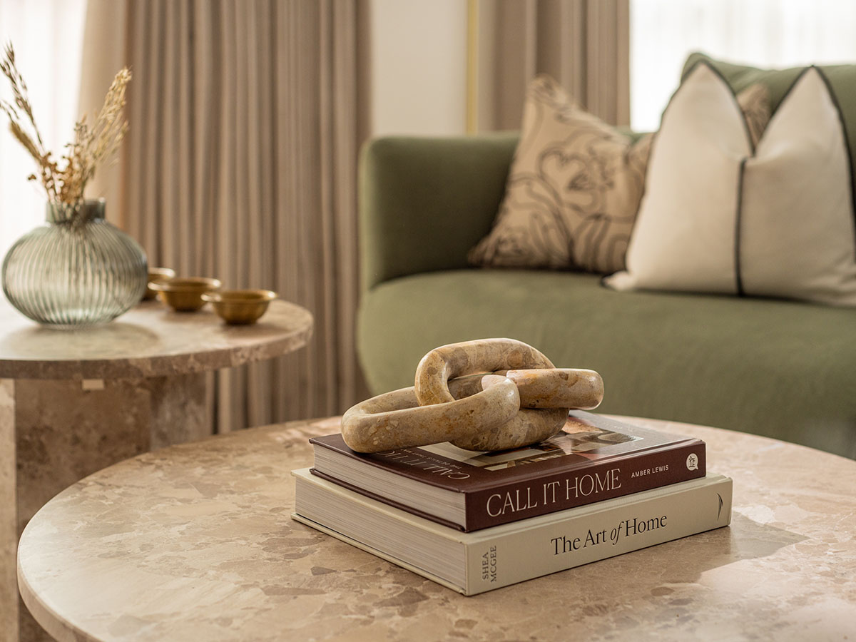 Close-up of luxury coffee table styling by InStyle Direct featuring circular marble tables topped with designer art books, a stone chain link sculpture, and a fluted glass vase, set against a backdrop of a green velvet sofa in Chelsea
