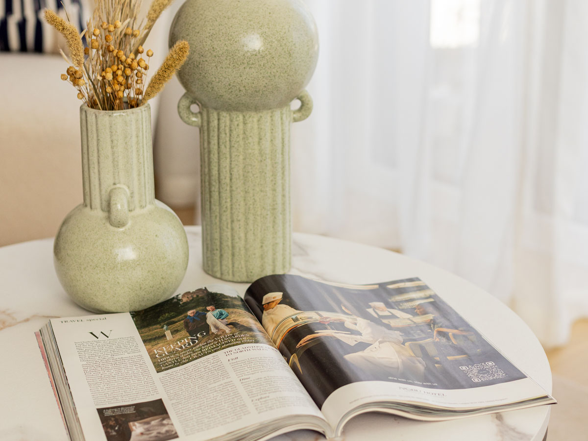 Close-up of coffee table styling by InStyle Direct featuring a pair of textured pale green ceramic vases with dried botanicals and an open lifestyle magazine on a white marble surface