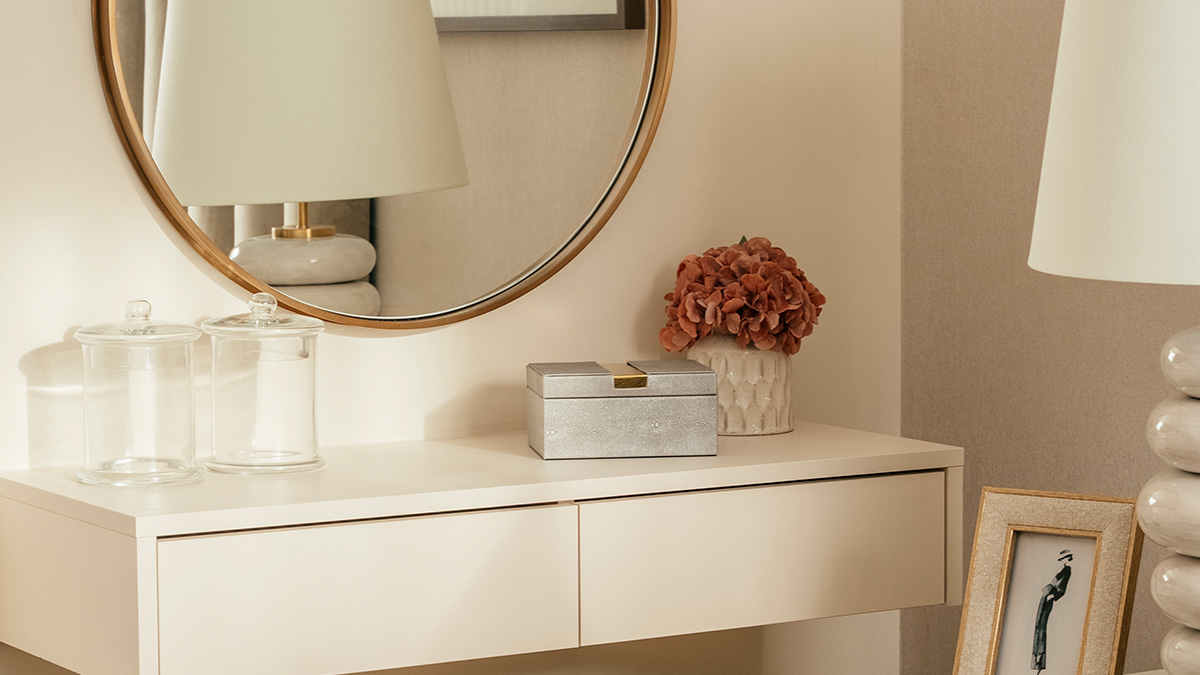 Modern minimalist vanity table with glass jars, a silver box, pink flowers, and a round mirror in a neutral-toned room.