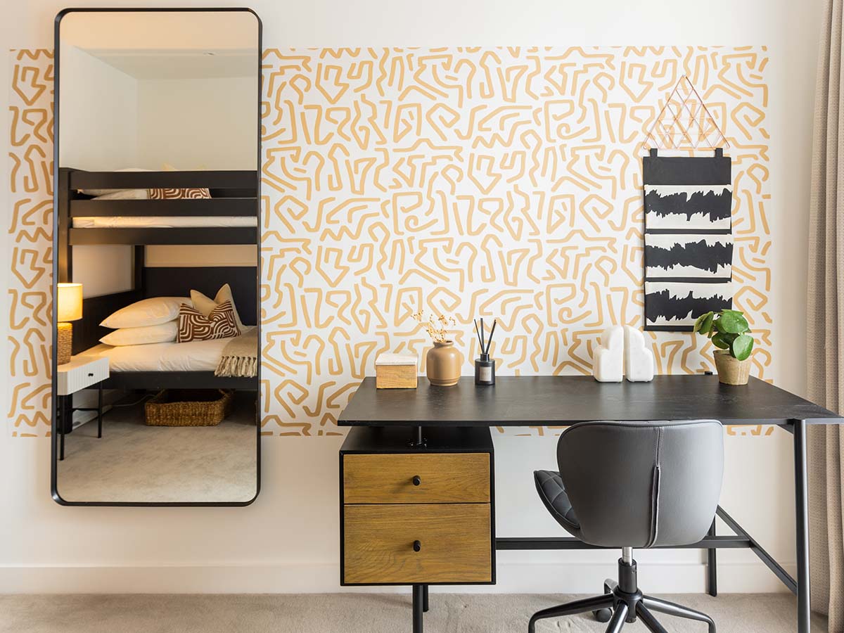A modern home office and bedroom space styled by InStyle Direct featuring a black minimalist desk with a grey swivel chair, a large black-framed leaning mirror reflecting a bunk bed, and a vibrant yellow geometric pattern accent wall in Wembley